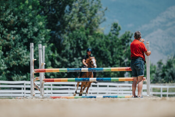 Horse riding instructor adjusts colorful jump bar while training a rider and horse outdoors during...