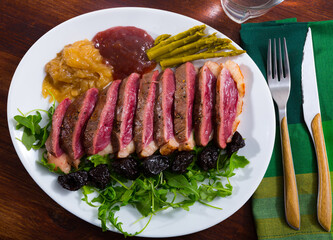 Top view of sliced fried duck magret with sauces, prune and vegetable garnish of pickled asparagus and fresh greens