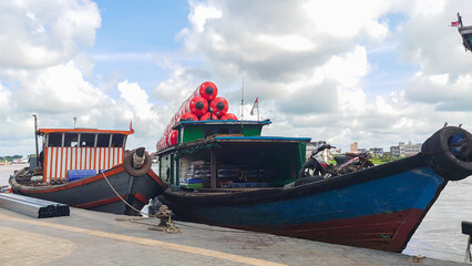 Two traditional boats are docked at a riverside, laden with goods and featuring vibrant colors...