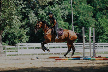A focused equestrian rider guides a horse over a jump in an outdoor training arena. The setting is...