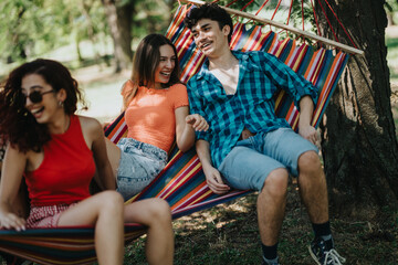 Fototapeta premium Three friends having fun and relaxing in a colorful hammock under a tree on a sunny day at the park.