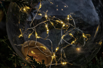 A closeup of warm glowing Christmas string lights inside a clear glass bulb. The Christmas ornament hangs on an evergreen tree for the holidays. The small festive twinkle LED lights have a yellow glow