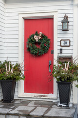 Naklejka premium The entrance to a white wooden house has a solid red door. A Christmas wreath hangs from the door. Two large black containers on either side are filled with fir, pine, and birch greenery.