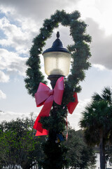 A lamppost light decorated for Christmas with a green wreath, tiny white lights, and a large red bow. The holiday light post is among trees. The red colored bow is made of cloth material at the bottom