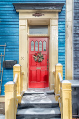 The exterior of a traditional wooden Colonial-style entrance to a house. The wall is bright blue, the door with small windows is red. There's a Christmas wreath hanging on the door. Snow on the ground