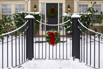 The entrance to a white house with a yellow wood and glass door. There's a black metal wrought iron fence and gate in the garden. A Christmas wreath hangs from it. The ground is covered in white snow.