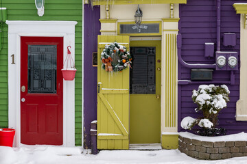 Exterior doors of houses from the sidewalk taken in winter with snow on the ground. The vibrant green and purple buildings have red and yellow doors. There's a Christmas wreath hanging on the door.