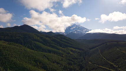 A scenic view of Mount Rainier with its snow-covered summit partially shrouded by clouds, surrounded by vibrant green forested hills.