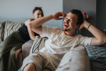 Fototapeta premium A cheerful man lounges on a comfortable sofa, smiling and enjoying a relaxed moment, while a friend sits in the background. Captures a friendly and laid-back atmosphere.