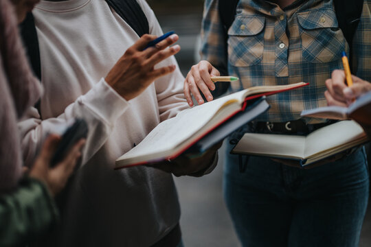 A close-up view of students engaged in a group discussion outdoors, using open books and notebooks. The scene conveys teamwork, learning, and academic collaboration.