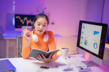 Young asian businesswoman wearing headphones around her neck working with computer and taking notes in notebook, studying trading charts displayed on computer screen