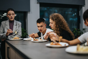 Students gather around a table, enjoying a meal together in an informal school setting. The atmosphere is relaxed, highlighting community and social interaction.