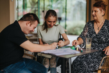 An elderly woman assists a male with Down syndrome and a girl in a wheelchair as they draw and...