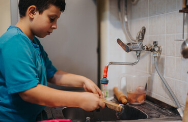 A young boy in a blue shirt washes dishes in a kitchen sink, emphasizing responsibility and cleanliness.