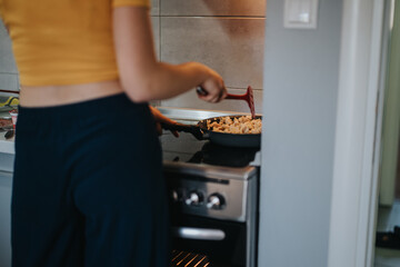 A person in a yellow top and dark pants cooking food in a frying pan on a stove in a modern kitchen.
