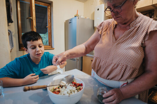An elderly woman and her grandson enjoy quality time in the kitchen, preparing a salad together. The cozy atmosphere reflects family bonding and the importance of sharing traditions across generations
