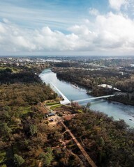 A breathtaking aerial view of a flowing river, modern Sundial Bridge, and lush parkland, surrounded by sprawling urban landscapes under a sky filled with soft clouds