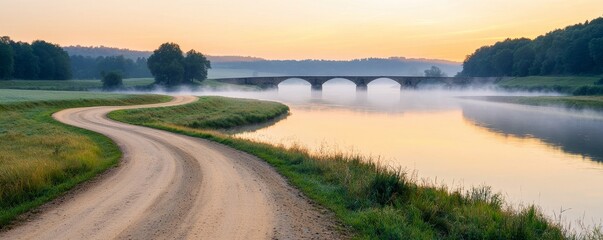 Fototapeta premium A serene river scene at sunrise, featuring a winding dirt path, lush greenery, and a misty atmosphere with a bridge spanning the calm waters.