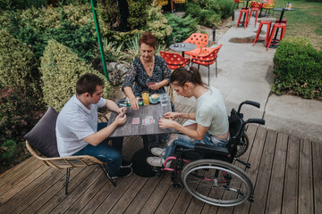 A boy with down syndrome, a girl in a wheelchair, and an older woman bond while playing cards outdoors. They enjoy a warm, sunny day together, having fun and building relationships.