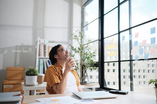 One Asian male freelance worker relaxes by drinking coffee and daydreaming about holiday after finishing deadline work in casual small business office, satisfied online startup jobs, E-commerce SME. - Powered by Adobe
