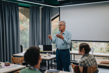 An elderly professor teaches a group of students in a bright, modern classroom. The image captures an interactive learning environment with focused students and an experienced educator.