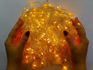 A woman's hands are holding a yellow glowing garland on a white background. The LED garland is yellow, orange in color. The lights of the diode garland.