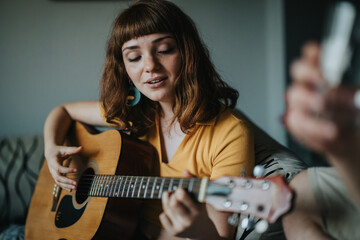 A young woman playing an acoustic guitar enjoys a peaceful moment in a cozy living room. The natural lighting adds warmth and relaxation to the scene.
