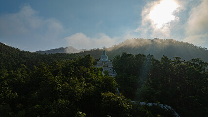 A serene aerial view of a large white Buddha statue perched on a forested hill, surrounded by lush greenery and overlooking a peaceful valley with distant villages. 