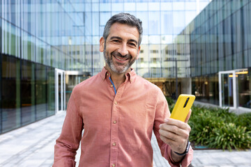 Mature happy man using mobile phone outdoors in the city looking at camera smiling.