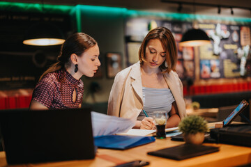 Two businesswomen engaged in a collaborative brainstorming session, analyzing documents and taking notes. The setting is a contemporary office environment during a late night team strategy meeting.