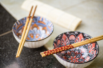 Two empty multi-colored unique patterned china bowls with chopsticks on top and sushi rolls on the base