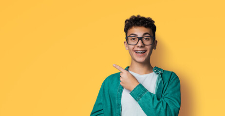 Happy black curly hair man in braces, brackets, eyewear, open mouth, wear glasses, green shirt advertise show point area for sale slogan text, isolate yellow background. Dental care ophthalmology ad.