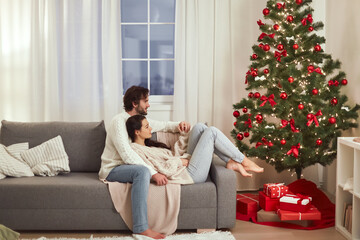 A couple relaxes together on a sofa in a cozy living room adorned with a Christmas tree and gifts....