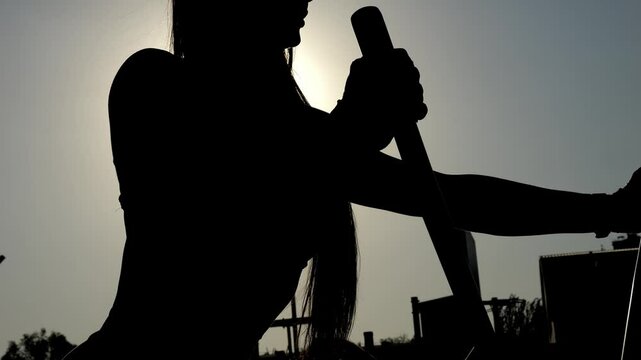 A close-up silhouette of a slender woman training on an outdoor orbitrek simulator. An athletic woman with long hair does cardio workout outdoors in summer in the setting sun.