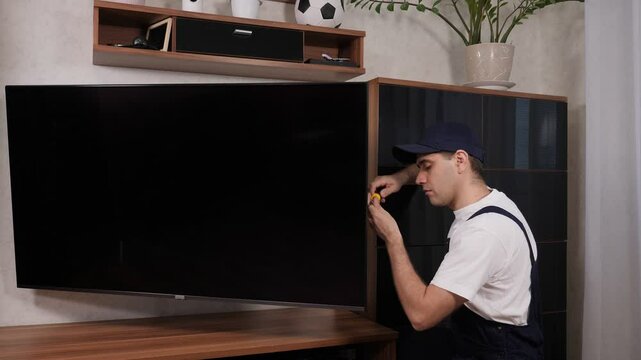 A male technician in blue overalls from customer service is repairing a TV at a customer's home. 