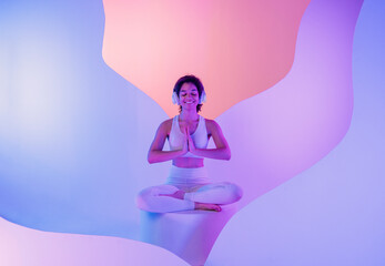 A young African in white sportswear sits in a meditation pose on round platform. A smiling black...