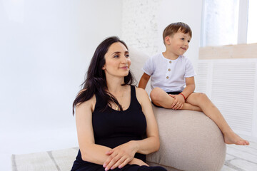 A young pregnant woman in a black dress is sitting with her son on the couch. Adorable mom and her little boy on the sofa.