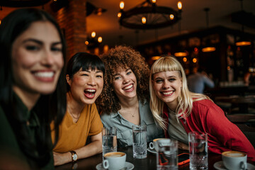 A young group of people sitting around a cafe table, taking a group selfie and drinking coffee.