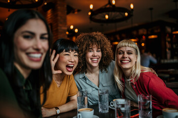 A young group of people sitting around a cafe table, taking a group selfie and drinking coffee.