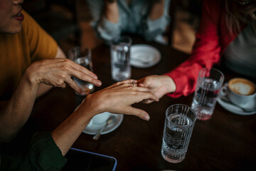 An unrecognizable woman shows her engagement ring to her female friends while drinking coffee in a café indoors.