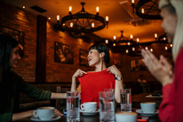 Carefree female friends have fun as they hang out together over coffee. Focus on a smiling Asian woman showing her new clothes