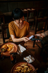 Mid aged Asian woman paying contactless using a credit card in the bar, with friends. A customer paying through credit card using contactless technology
