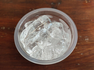 A close-up view of crystal-clear ice cubes in a transparent plastic bowl placed on a wooden table. Perfect for beverage concepts, summer themes, and refreshing visuals.