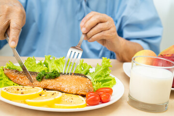 Asian senior woman patient eating Salmon steak breakfast with vegetable healthy food while sitting and hungry on bed in hospital.