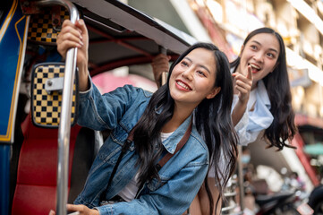 Two excited Asian girl tourists enjoy a ride in a tuk-tuk in Thailand, pointing at the view.