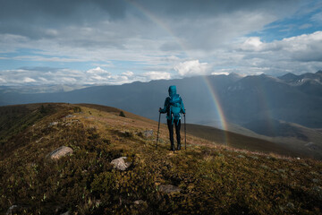 Woman backpacker hiking on high altitude mountain top after the rain, double rainbow in the background