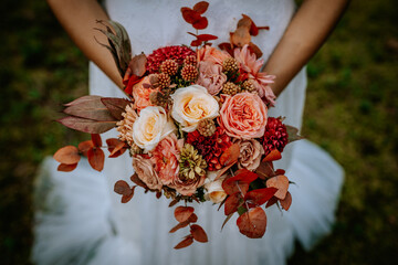 Close-up of a bride holding a beautiful bouquet of autumn-themed flowers in warm tones of peach,...