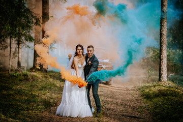 Bride and groom celebrate with orange and blue smoke bombs in a forest, creating a colorful and...