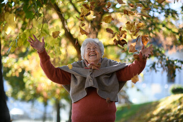 Portrait of elderly woman on walk in the park enjoying warm autumn day, throwing colorful leaves.