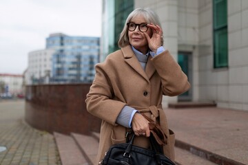 Stylish elderly woman adjusting her glasses while walking in a city during the afternoon in a warm coat and carrying a handbag